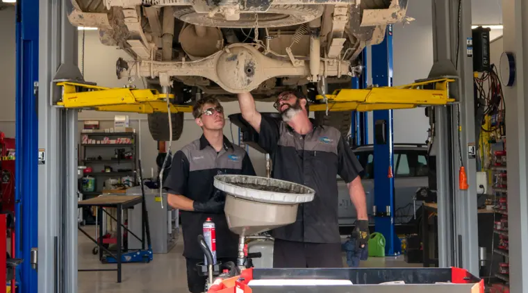 Two men wearing safety glasses and gloves are working under a vehicle in an automotive workshop.